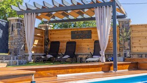 Area piscina con pergola in legno, pareti in pietra e poltrone relax.