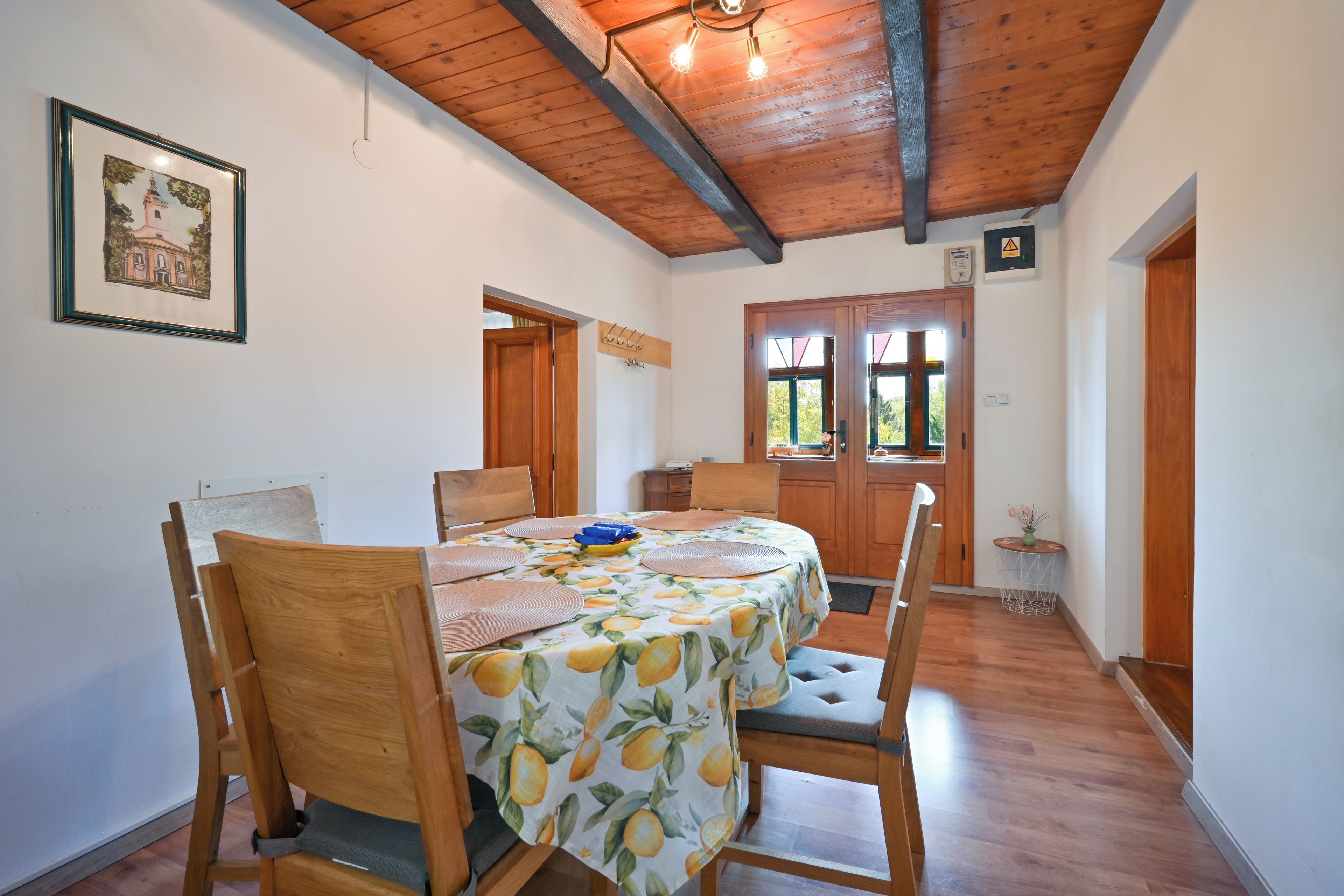 A dining area with a table set for four, wooden chairs, a ceiling with exposed beams, and a framed picture on the wall.