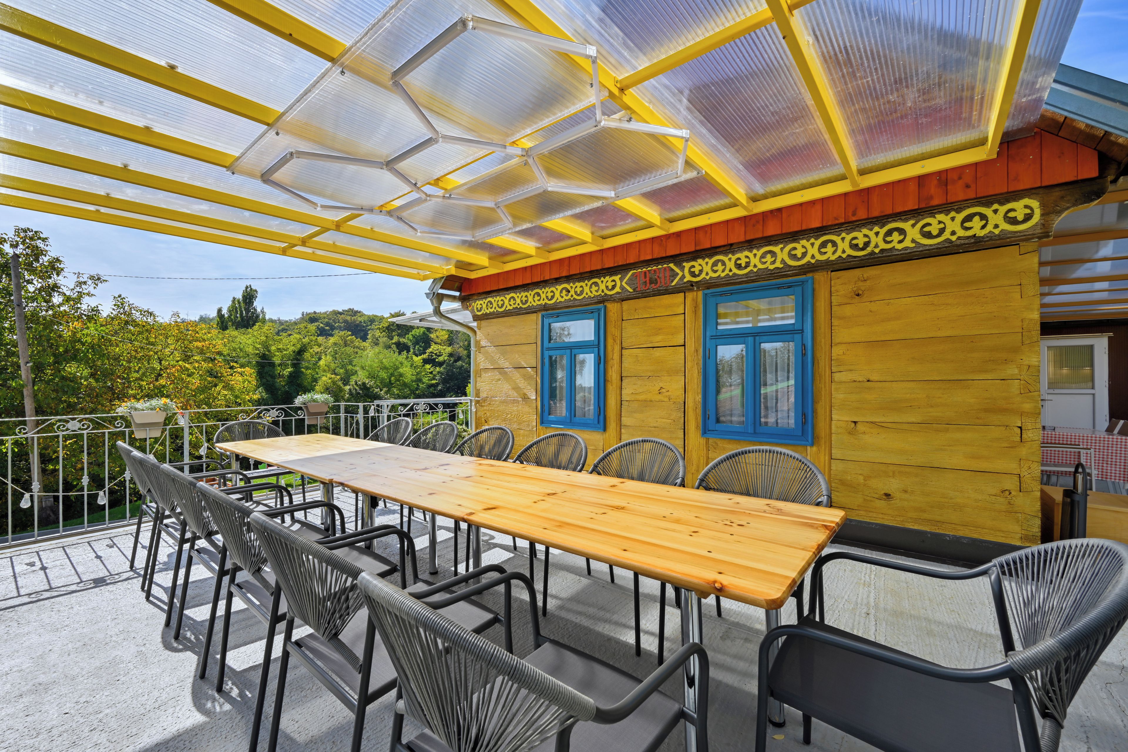 Un espace de restauration en plein air avec une longue table en bois et des chaises en métal, un mur extérieur jaune et rouge, et une structure de toit en verre.
