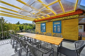 An outdoor dining area with a long wooden table and metal chairs, a yellow and red exterior wall, and a glass roof structure.