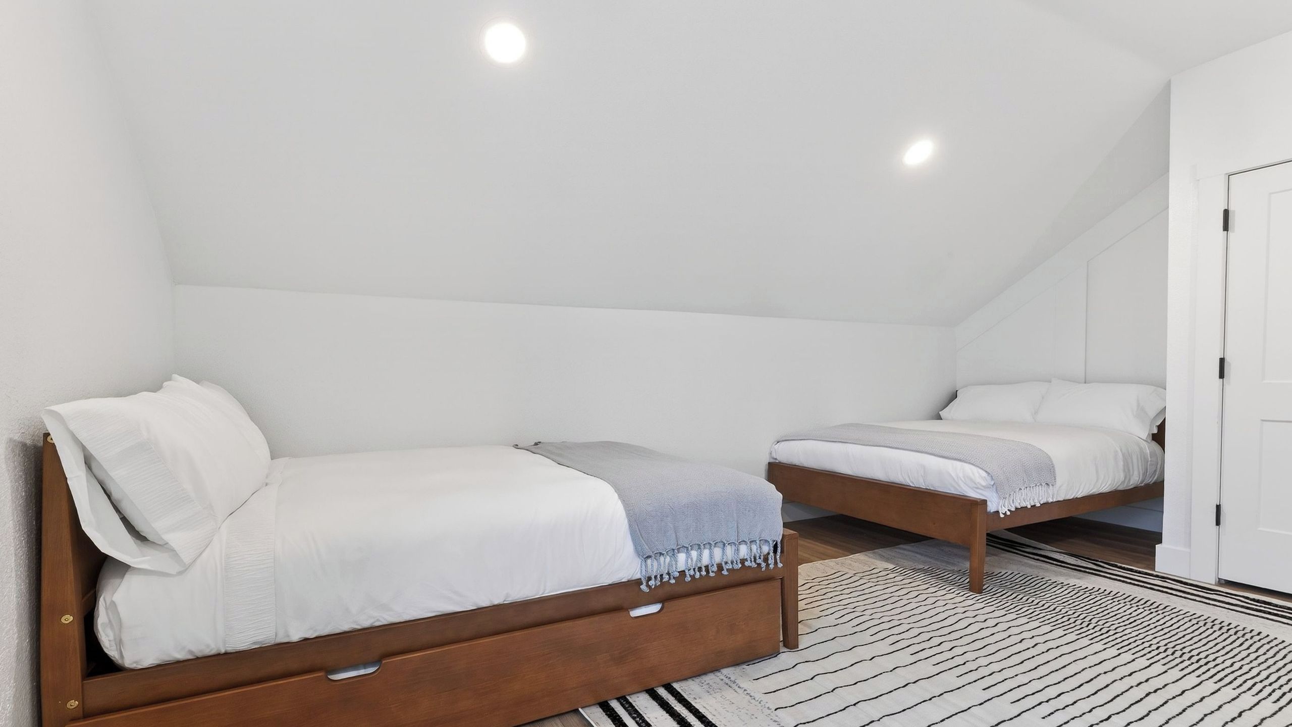 Quarto com duas camas de solteiro, cabeceira de madeira, tapete listrado e porta branca.