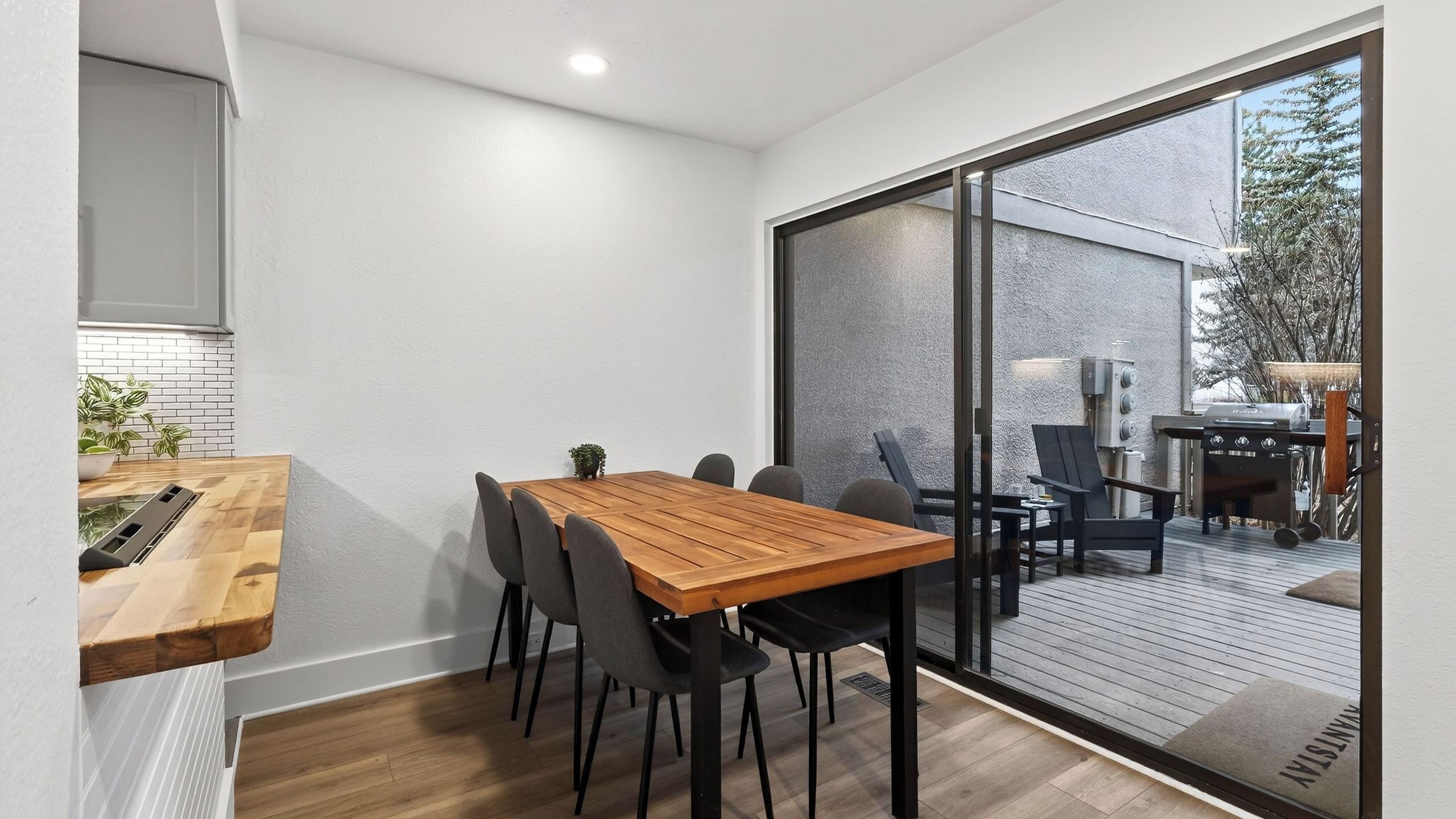 A modern dining area with a wooden table and chairs, a small plant, and a view of the outdoors through a glass door.