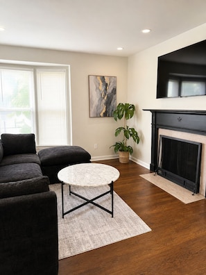 A living room with a sectional sofa, a round coffee table, a fireplace, a large abstract painting, and a potted plant.