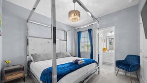 A modern bedroom with a four-poster bed, a bedside table, a chair, and a bathroom visible through an open door. - Ultimate Disney Getaway | 12BR Bella Vida 30+ (Kissimmee)