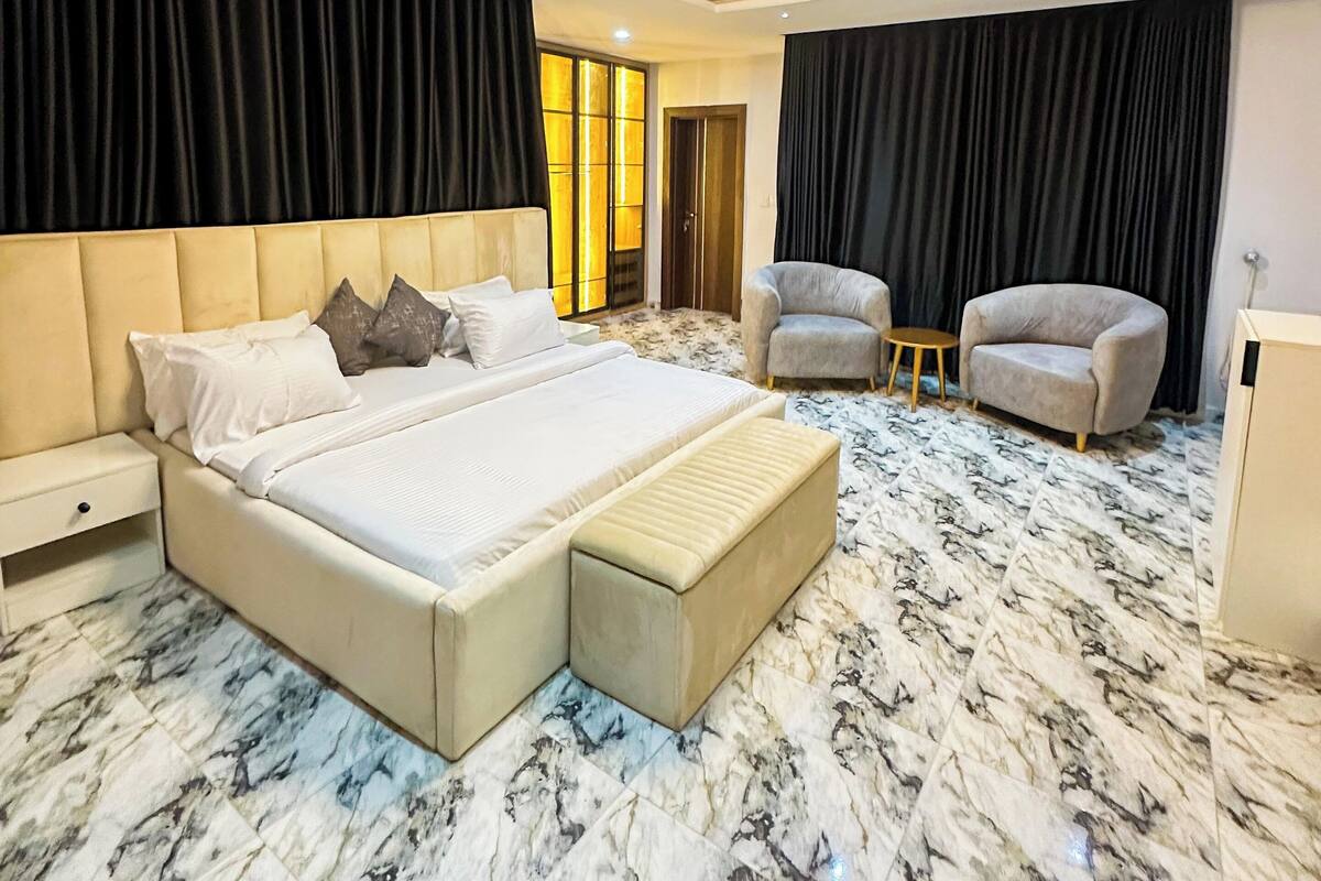 A spacious bedroom with a large bed, two armchairs, a small table, and a wardrobe.