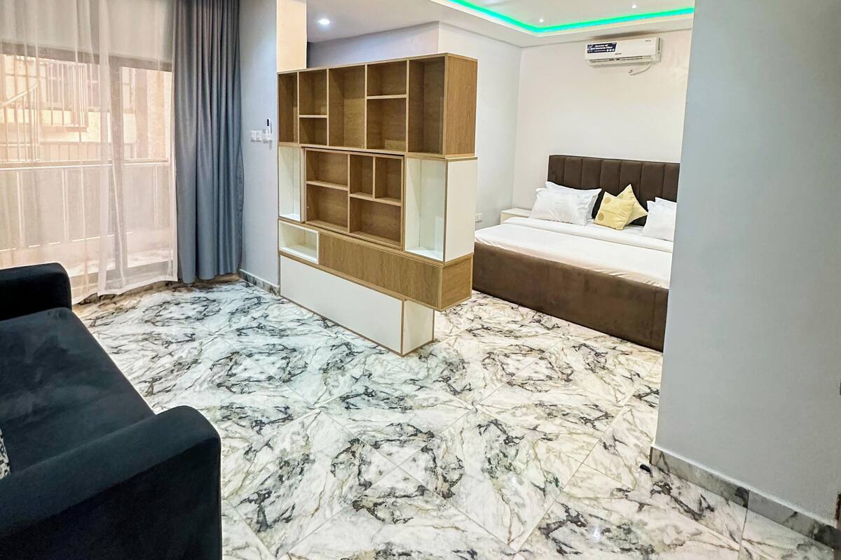 A modern hotel room with a bed, a sofa, a built-in wooden shelf unit, and a marble-patterned floor.
