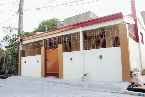 Un edificio de dos pisos con techo rojo, paredes blancas y entrada protegida por una reja.