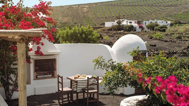 Eine Terrasse mit Tisch und Stühlen, eine weiße Wand mit Fenster und ein Blick auf einen Weinberg im Hintergrund.