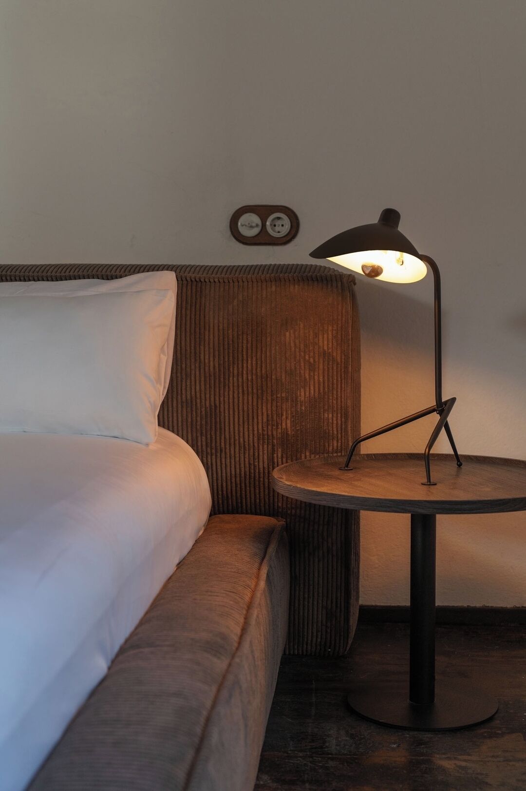 Habitación de hotel con cama, una mesita redonda con lámpara y un enchufe de pared.