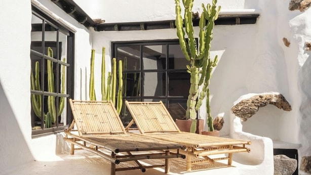 Zona relax all'aperto illuminata dal sole, con due poltrone in bambù, circondata da cactus e un edificio bianco con base in pietra.