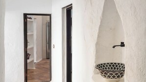 Un corridoio con pavimento piastrellato, una parete bianca con uno scaffale su cui ci sono una ciotola e una pianta, e una porta che dà su un'altra stanza.