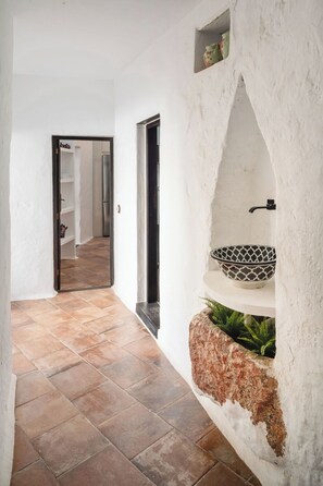 A hallway with a tiled floor, a white wall with a shelf holding a bowl and a plant, and a door leading to another room. - Canaryislandshost I La Casa del Doctor (Teguise)