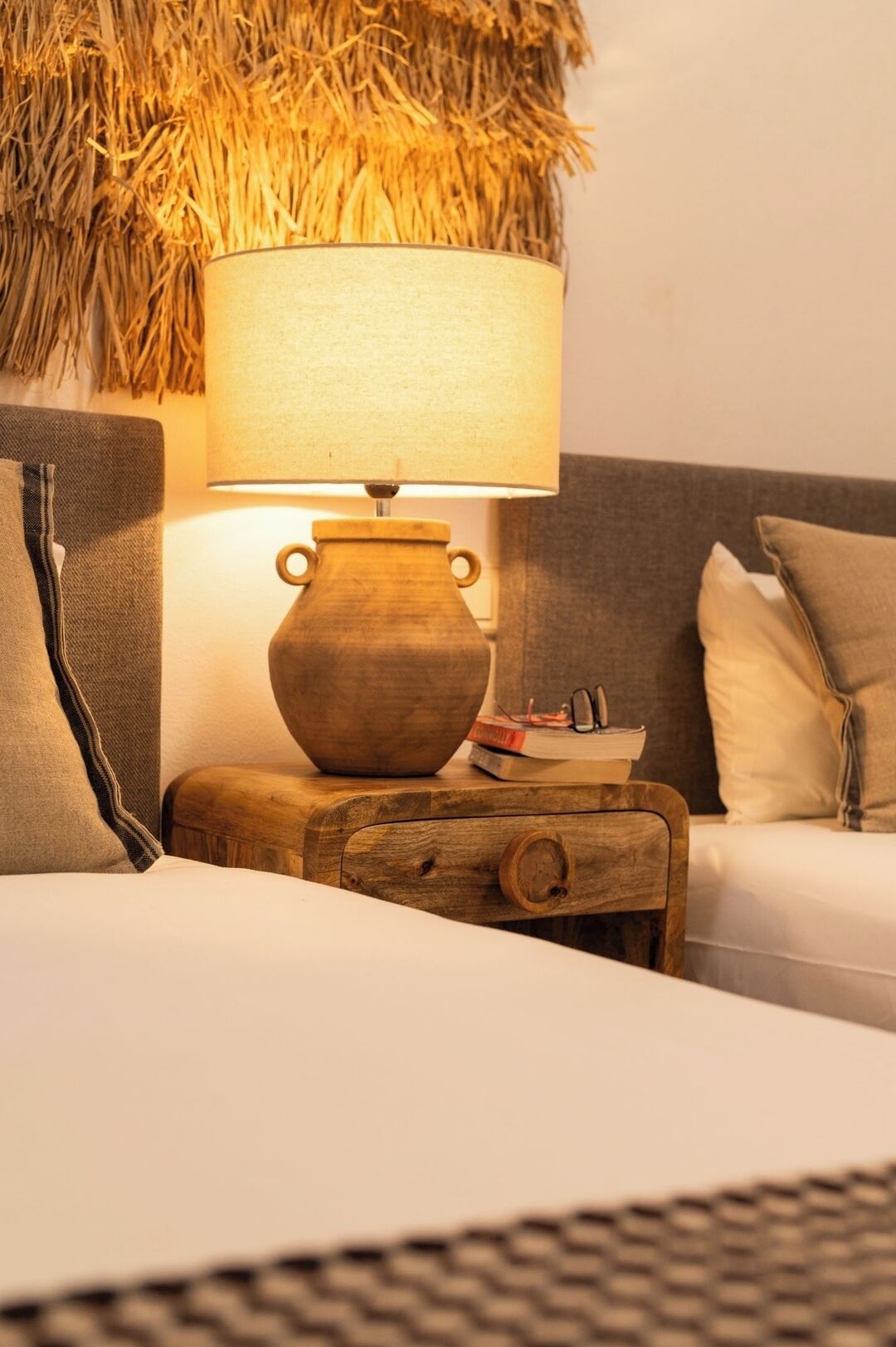 Una cama con una mesita de noche de madera a su lado, una lámpara sobre la mesita y una pared con textura que recuerda al paja.