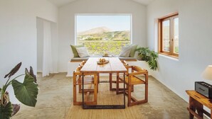 Ein Essbereich mit Tisch und Stühlen, Blick auf eine Landschaft durch ein großes Fenster und eine Pflanze auf der rechten Seite.
