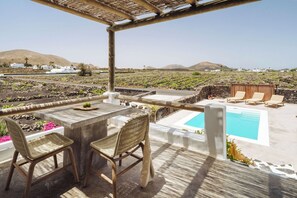 Terraço com piscina, duas cadeiras de vime, uma mesa e vista para paisagem rural com edifícios brancos e montanhas.