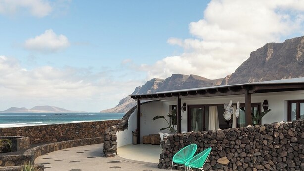 Une propriété située sur la côte, dotée d’un mur en pierre, d’une terrasse avec des chaises et offrant une vue sur l’océan et les montagnes.