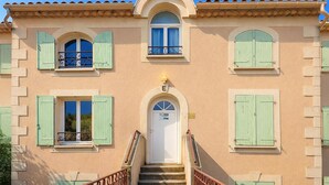 Un edificio de dos pisos con una entrada central, contraventanas verdes y una escalera que lleva al segundo piso.