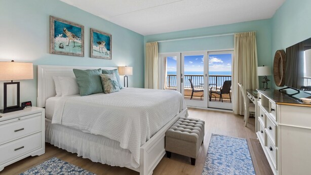 A bedroom with a bed, bedside tables, a dresser, a bench, a balcony with a view, and artwork on the wall. - Oceanfront Condo in Amelia Island Plantation with Pool (Fernandina Beach)