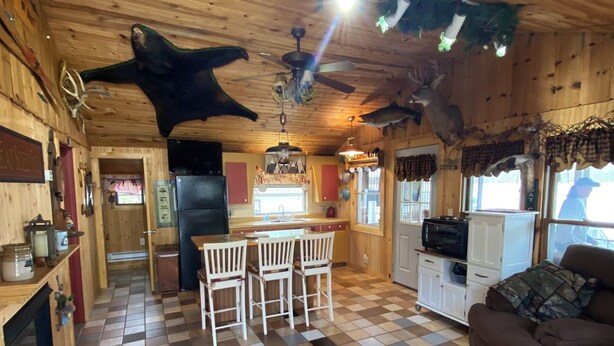 A rustic cabin interior with a checkered floor, wooden walls, a kitchen area, and a seating area with a couch and chairs.