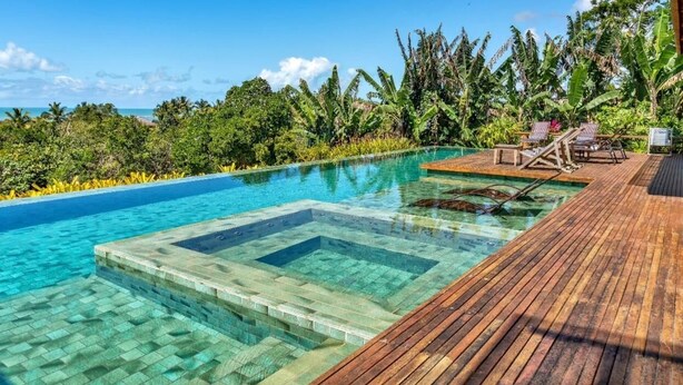 Ein klarer Swimmingpool mit Holzterrasse und Liegestühlen, umgeben von tropischer Vegetation und Blick aufs Meer.