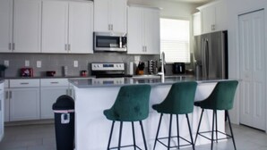 Une cuisine moderne avec des armoires blanches, un réfrigérateur en acier inoxydable, un four à micro-ondes et une cuisinière. On trouve trois tabourets de bar verts près de l’îlot central.