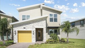 Une maison à deux étages avec une façade en pierre, une porte de garage jaune et un petit jardin devant, agrémenté de palmiers.