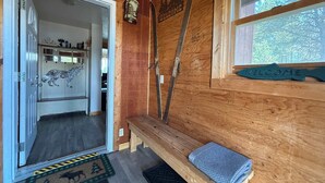 L’intérieur d’un chalet en bois, avec un banc, une pancarte de bienvenue et une porte donnant sur une pièce dont un mur est décoré d’une fresque représentant un dinosaure.