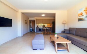 Uma sala de estar moderna com um sofá cinza, uma mesa de centro de madeira e um banquinho cinza. Há uma televisão montada na parede e um quadro sobre o sofá.