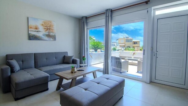 A modern living room with a grey sofa set, a wooden coffee table, and a grey bench. A painting of trees is on the wall, and there is a sliding glass door leading to an outdoor area with a view of buildings and greenery. - Vakakis Family Luxury Studios (Kantanos-Selino)