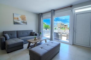 Un salon moderne avec un ensemble de canapés gris, une table basse en bois et un banc gris. Un tableau représentant des arbres est accroché au mur, et une porte-fenêtre coulissante donne accès à un espace extérieur d’où l’on peut admirer des bâtiments et de la verdure.