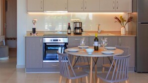 A modern kitchen with a built-in oven, a coffee maker, and a dining area with a round table and chairs. - Vakakis Family Luxury Studios (Kantanos-Selino)
