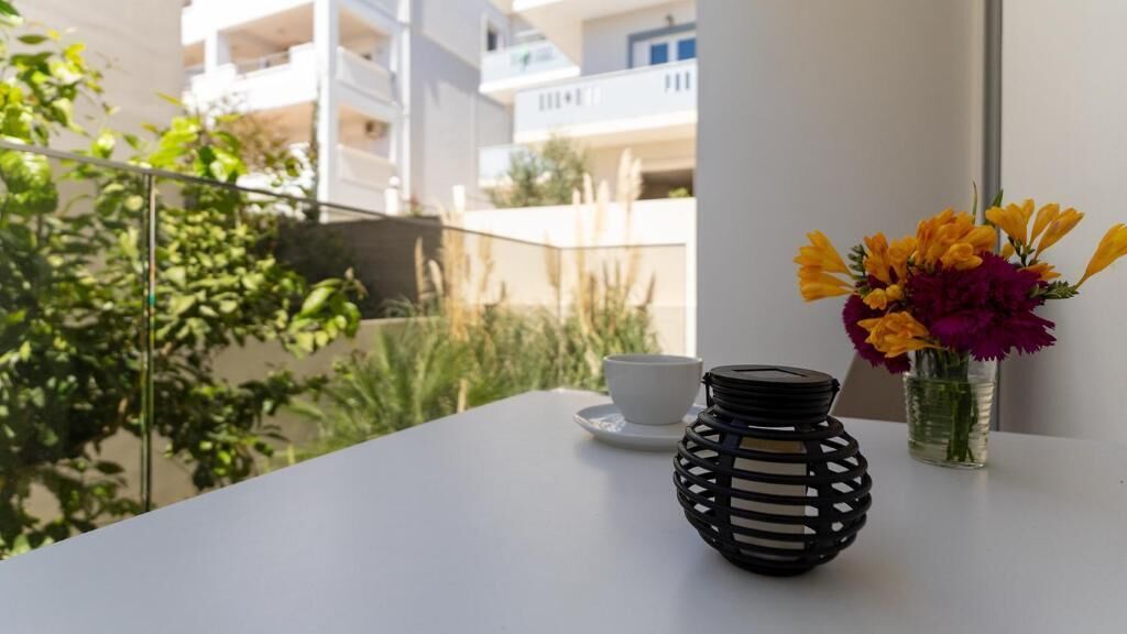 Un balcón con una mesa blanca, una lámpara tejida negra y un florero con flores.