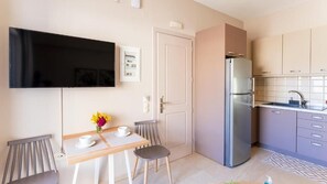 A compact kitchen with a refrigerator, a dining table with chairs, a flat-screen TV mounted on the wall, and a small window. - Vakakis Family Luxury Studios (Kantanos-Selino)