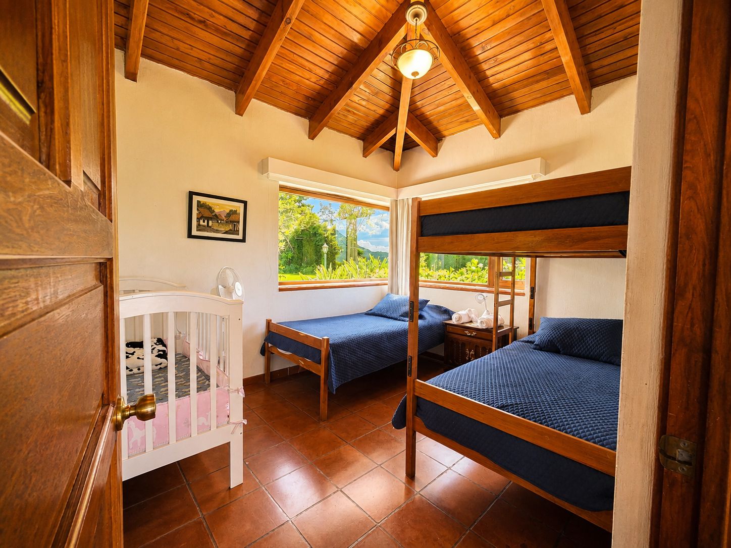 A room with a high wooden ceiling, two bunk beds, a crib, and a window with a view of greenery.