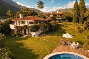 A two-story house with a swimming pool, surrounded by lush greenery and mountains.