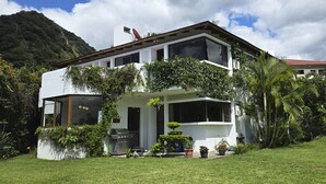 Una casa moderna de dos pisos con techo plano, rodeada de exuberante vegetación y con montañas al fondo.