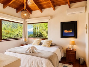 A bedroom with a large window, a bed with pillows, a nightstand with a lamp, and a framed picture on the wall.