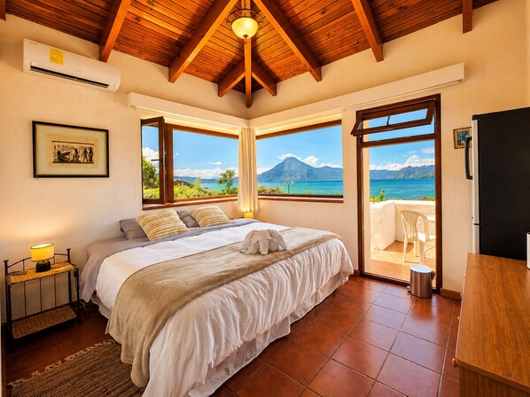 Un dormitorio con cama, mesita de noche, lámpara y ventana con vistas al mar y las montañas.