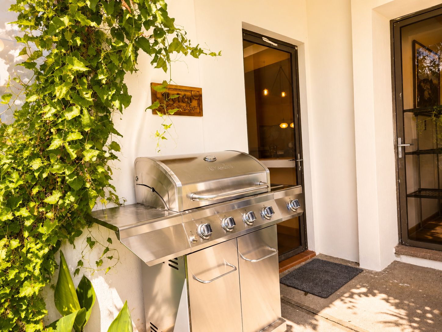 An outdoor grill with a stainless steel finish, positioned against a wall with a window and door.
