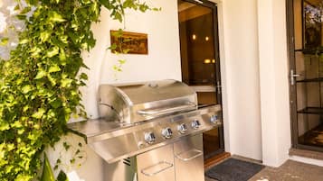 An outdoor grill with a stainless steel finish, positioned against a wall with a window and door.