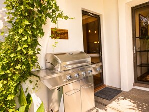 An outdoor grill with a stainless steel finish, positioned against a wall with a window and door.