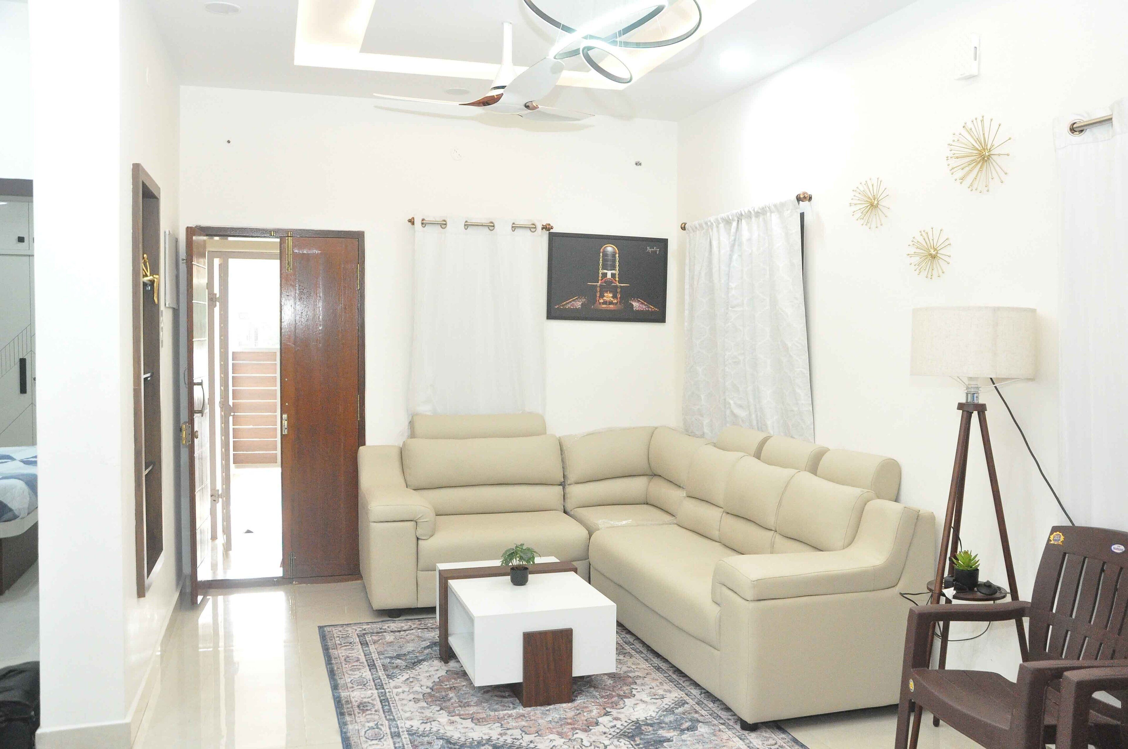 A living room with a beige sectional sofa, a small white coffee table with a plant, a floor lamp, and a wooden chair.