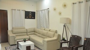 Una sala de estar con un sofá seccional color beige, una pequeña mesa de centro blanca, un sillón marrón oscuro, una lámpara de pie y un tapete estampado.