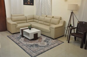 Un canapé d’angle beige, une petite table d’appoint blanche, une chaise en bois foncé, un lampadaire et un tapis à motifs dans un salon.