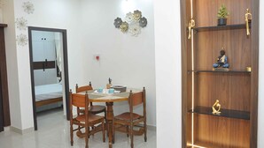 Una habitación con una mesa de comedor de madera, cuatro sillas, una cama con espejo, decoración en la pared y un estante de madera con una estatua.