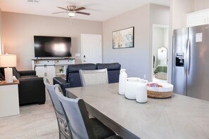 Ein modernes Wohnzimmer mit Flachbildfernseher, Deckenventilator, Esstisch mit Stühlen, einer Couch und einem Kühlschrank.