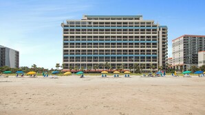 Un hôtel à plusieurs étages offrant une vue sur la plage, doté de nombreux balcons et d’une plage de sable aménagée avec des parasols et des chaises longues.