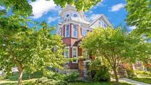 Une maison de style victorien, dotée d’une tour imposante, entourée d’une végétation luxuriante et d’une pelouse bien entretenue.