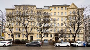 En bygning med flere etager, gul facade, parkerede biler og træer uden blade foran.