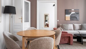En moderne stue med et rundt træbord, grå polstrede stole, en rød skammel, en grå sofa med mønstrede hynder og et sort sofabord, samt en gulvlampe.